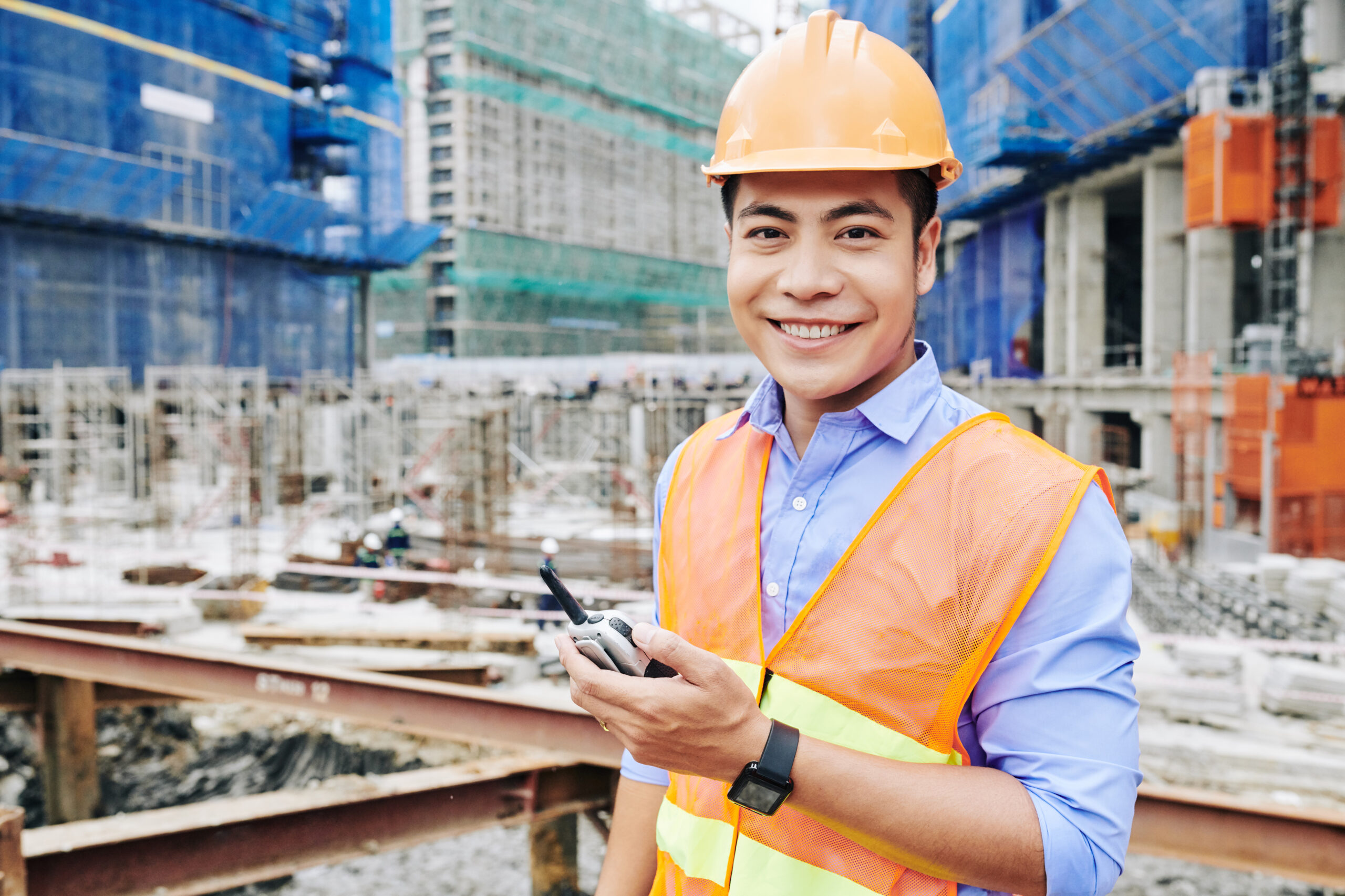 Portrait of cheerful Vietnamese construction engineer standing with portable radio communicator and smiling at camera